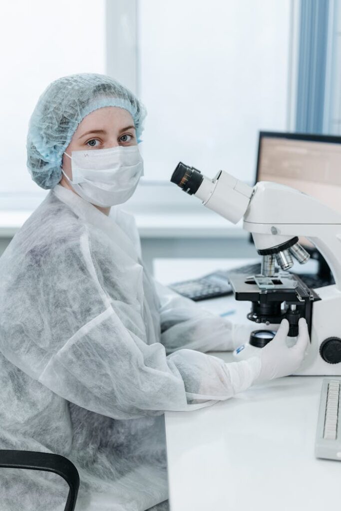 A woman scientist using a microscope in a lab, wearing protective gear for research.